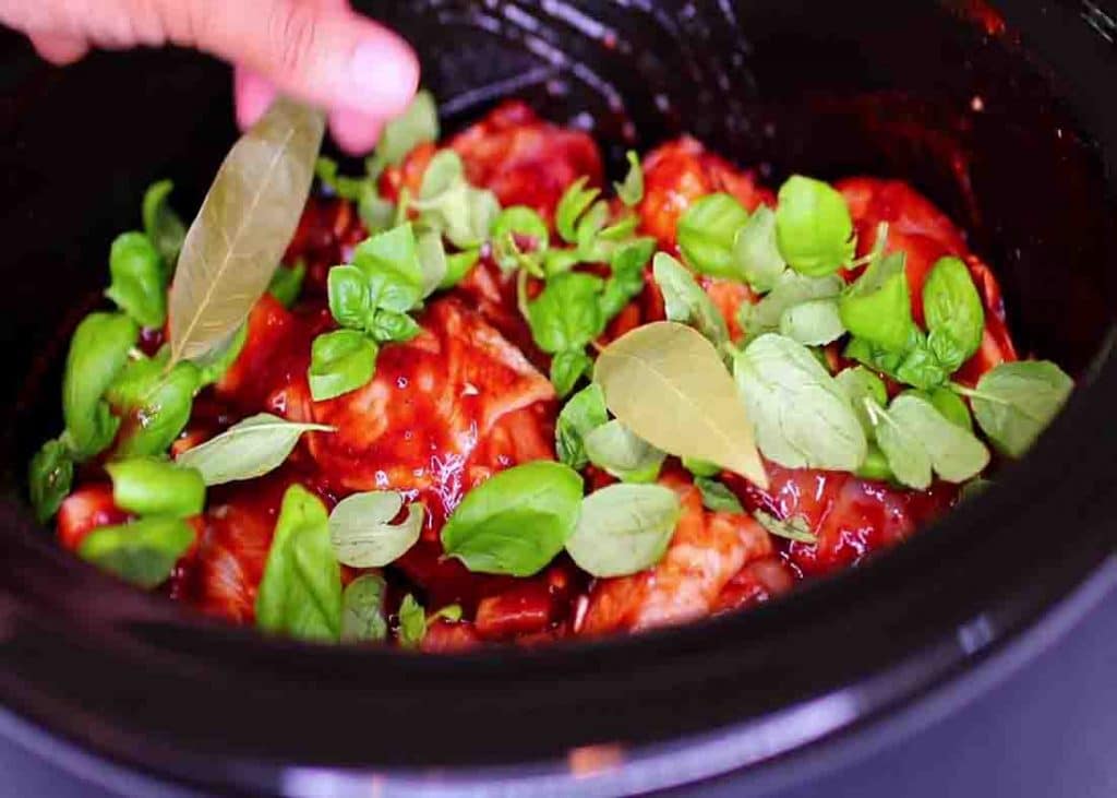 Adding the fresh basil leaves and dried bay leaves to the slow cooker with the chicken drumsticks
