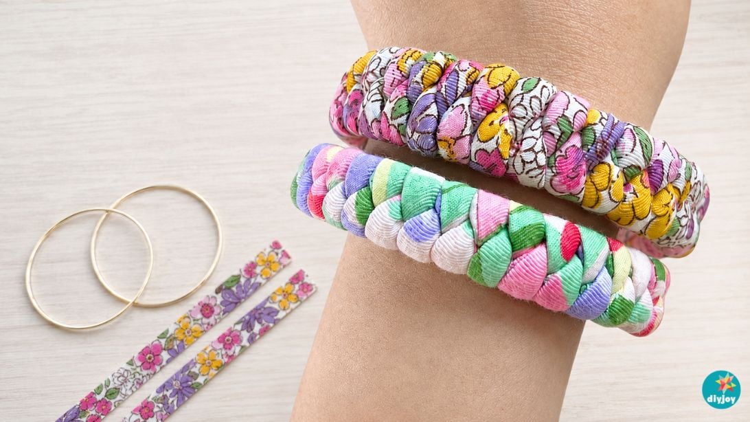 A wide braided fabric cuff bracelet in mixed prints with a small charm at the clasp, lying on a plain background