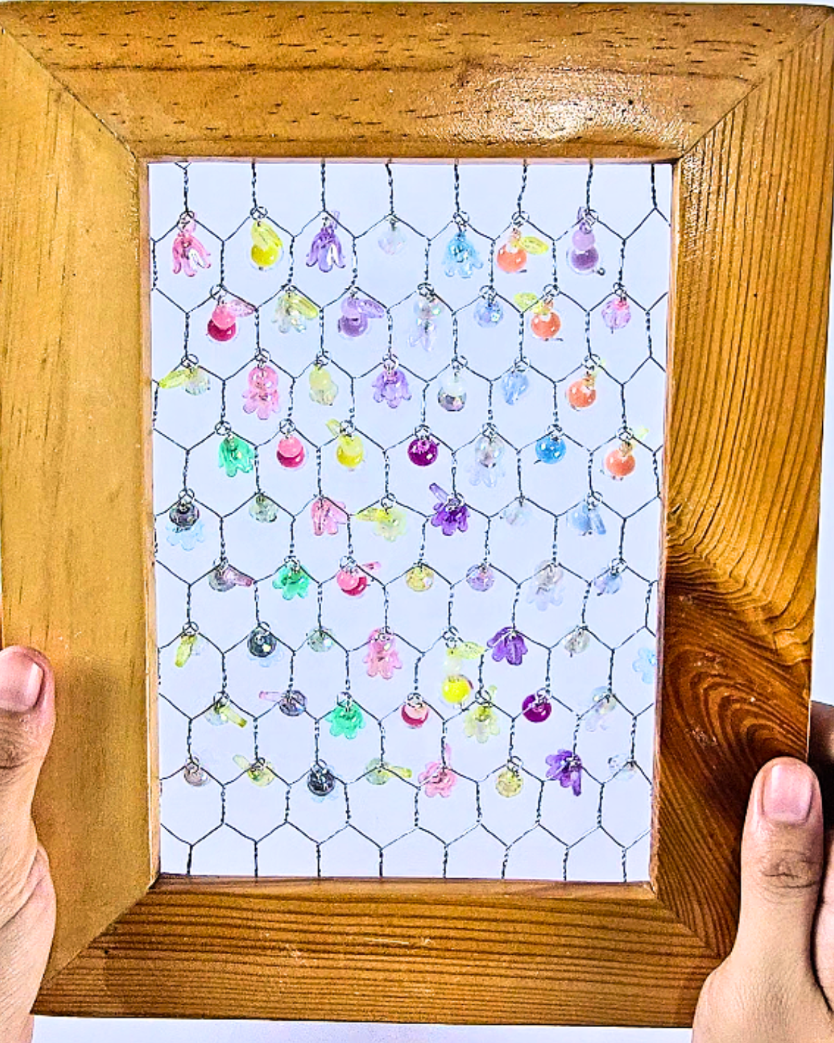 Hands holding a finished picture frame suncatcher with beads hanging on chicken wire.