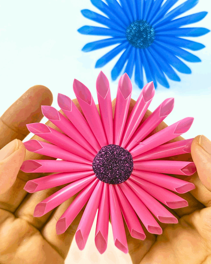 Holding a pink cocktail straw flower while showing the completed result, with a blue flower placed on the table.
