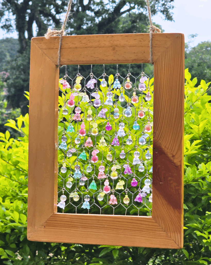 Picture frame suncatcher with beads hanging on a tree branch, plants in the background.