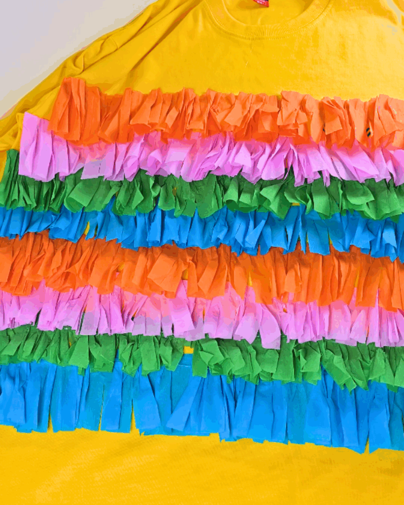 Last Minute Halloween Costume - the finished yellow shirt laid flat, covered edge to edge in repeating rows of orange, pink, green and blue crepe paper fringe