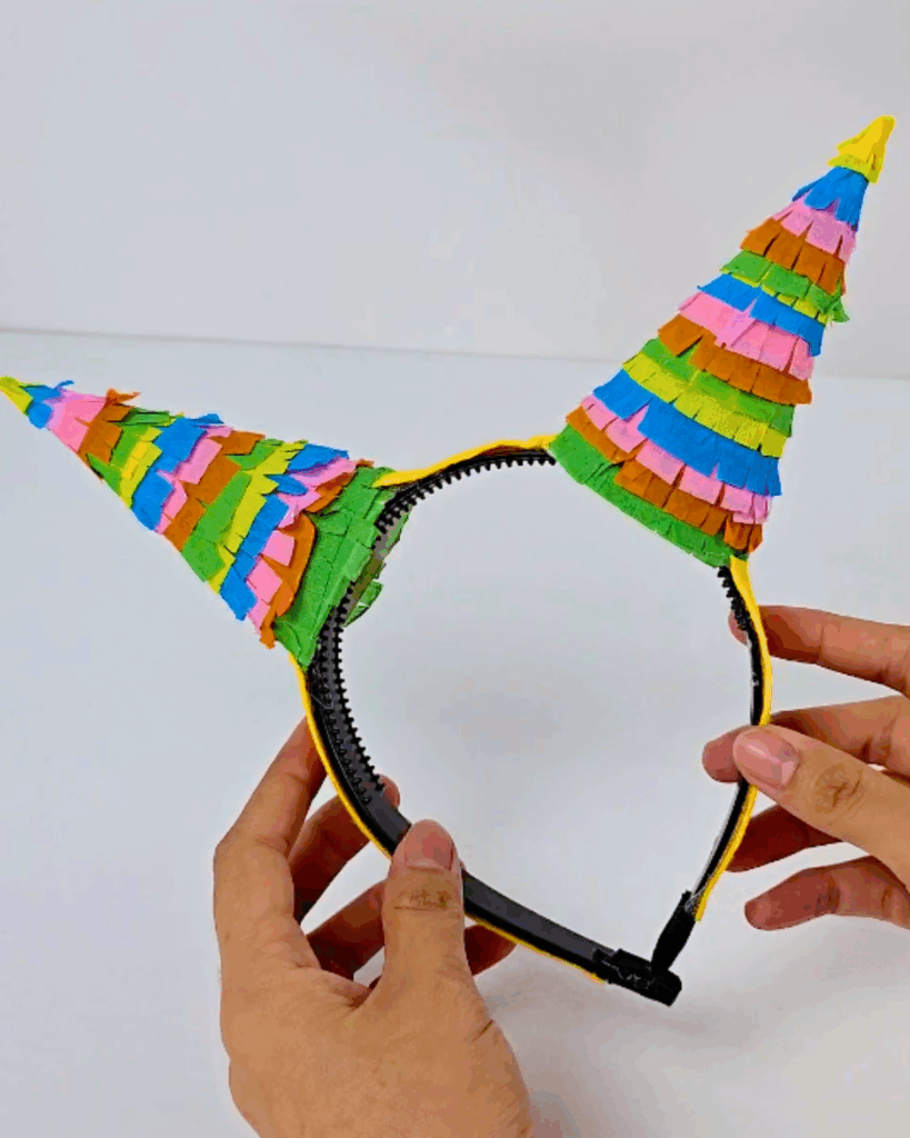 DIY Costume Accessories - a finished black headband held in two hands with two fringed cone horns attached, both covered in layered green, orange, pink and blue crepe paper