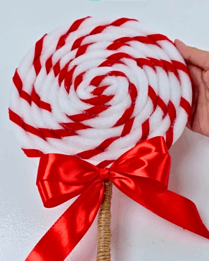 Candy Costume - the finished giant red and white swirl lollipop prop held up against a white wall, mounted on a jute twine wrapped stick with a red satin ribbon bow tied where the swirl meets the stick