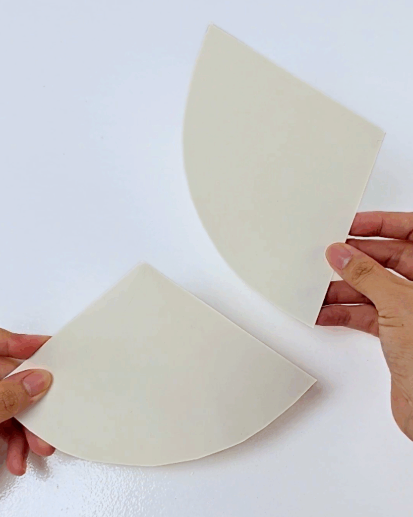 Cardboard Crafts - two hands holding two cream cardboard pieces cut into right angle triangles on a white surface, the flat shapes that become the headdress cones