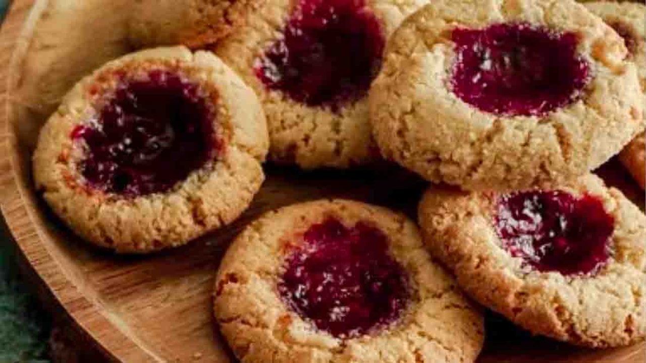 Vegan almond flour thumbprint cookies filled with dark red fruit jam, piled together on a round wooden board