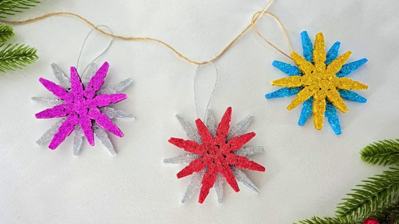 Three clothespin snowflake ornaments glittered in pink, red and gold, hung on twine beside a sprig of green pine