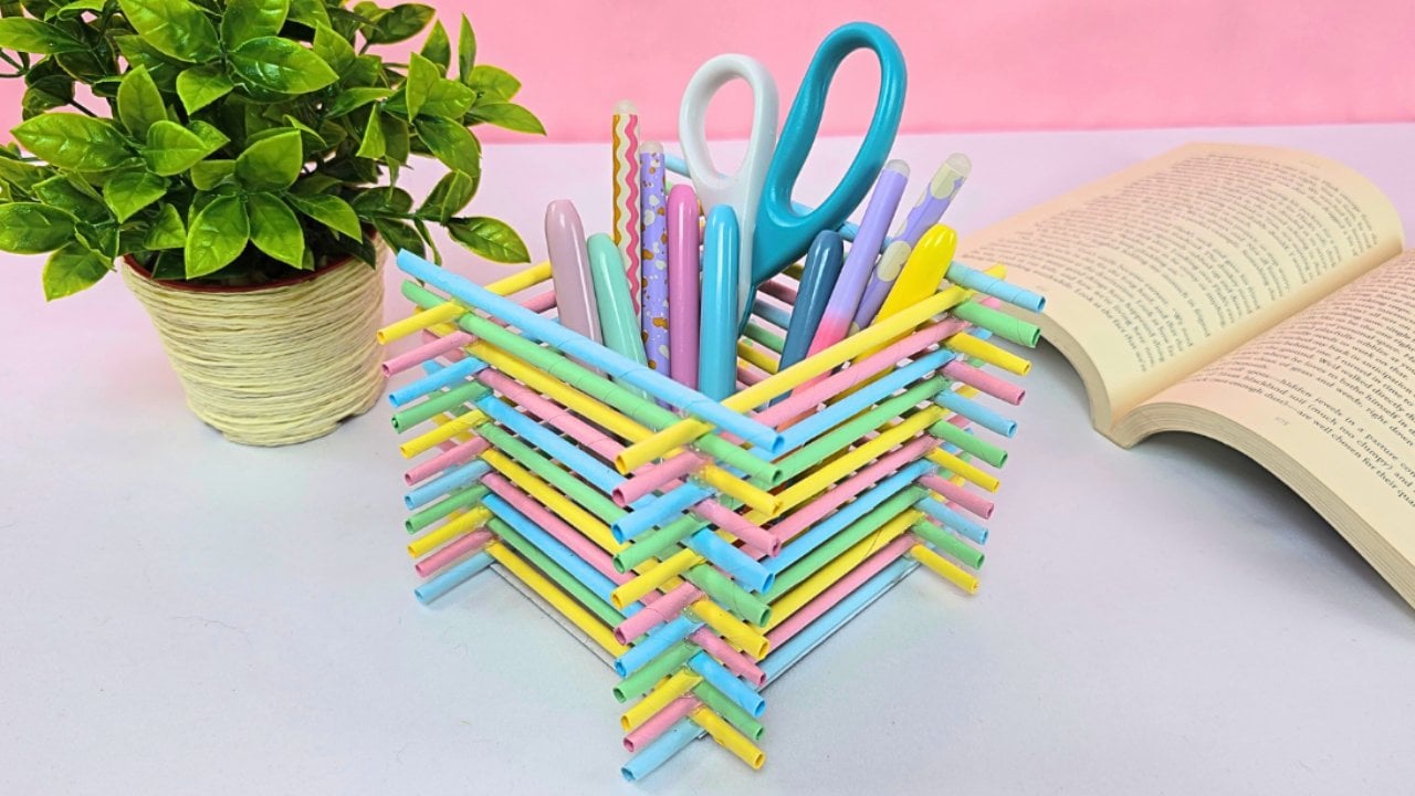 Pen holder built from pastel paper tubes stacked in a lattice, holding pens and scissors beside a plant