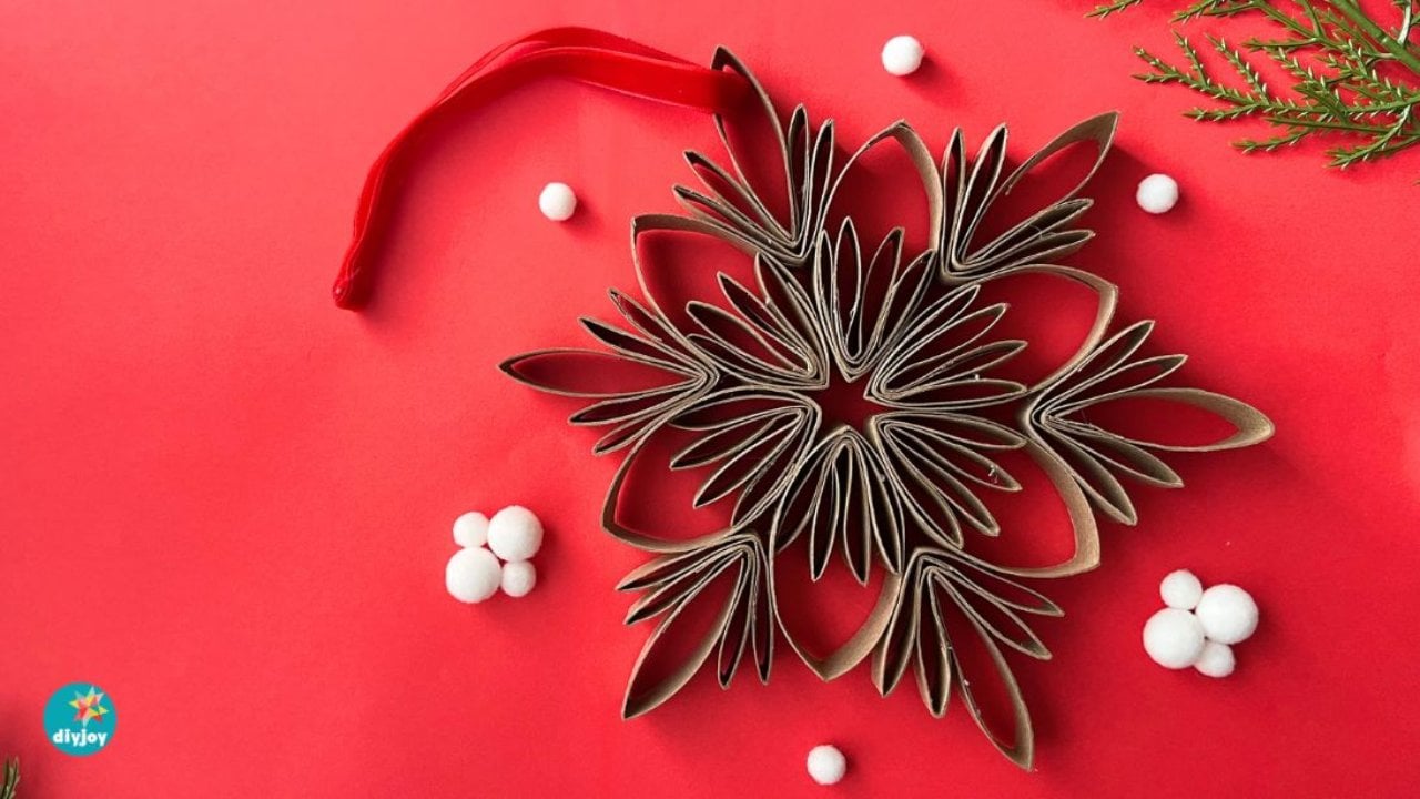 Finished snowflake ornament built from cut and folded cardboard tubes on red, with white pom poms and a red ribbon