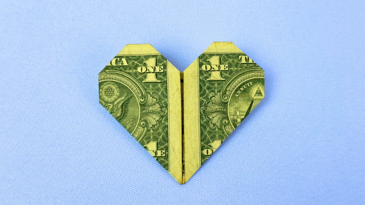 A neatly folded origami heart made from a dollar bill on a table, a creative money gift idea for him