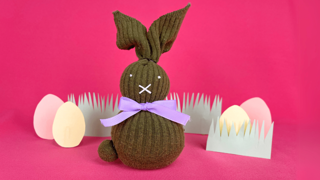 Sock bunny made from a brown sock filled with rice, with tall ears, a lilac ribbon bow and a stitched face by pastel eggs