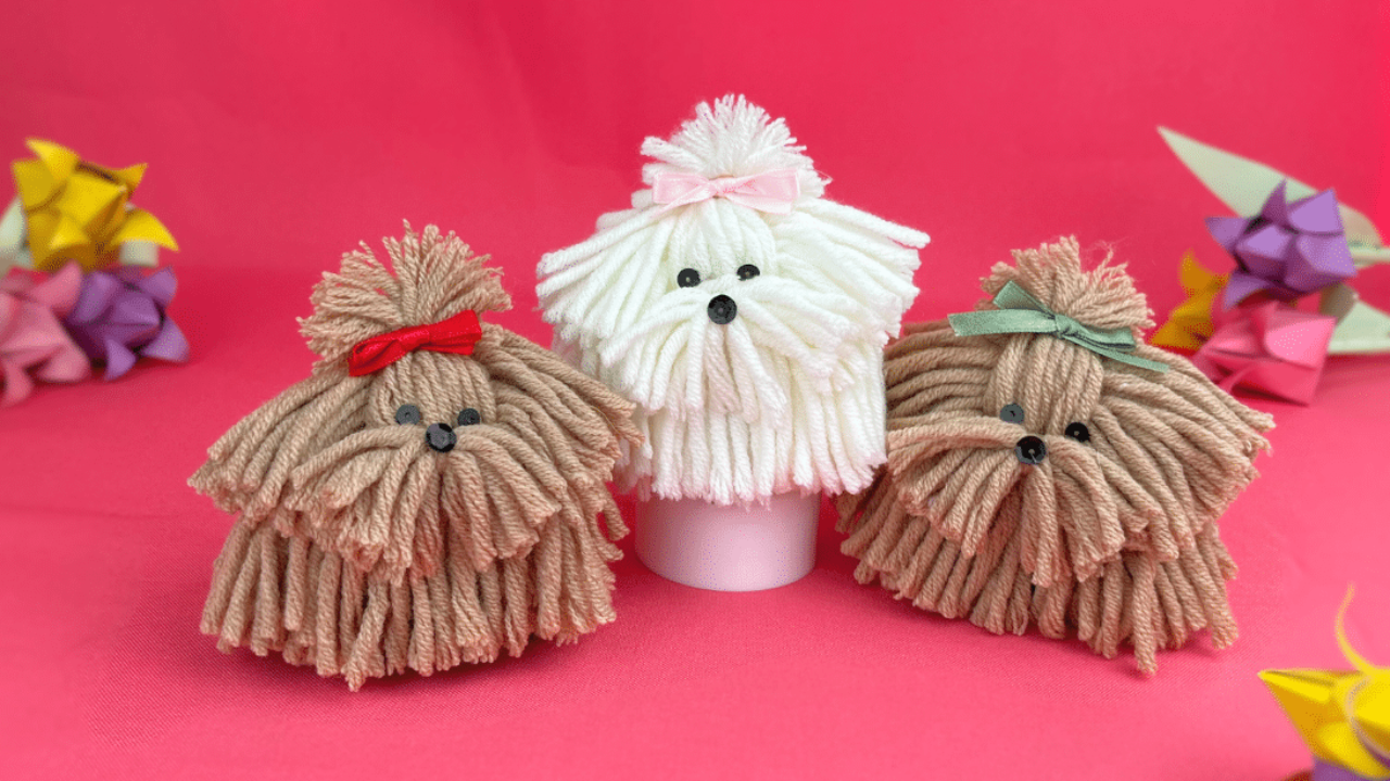 How to Make a Yarn Dog, Pom Pom Dog Craft Three yarn dogs with ribbon bows and bead eyes, two tan and one cream, on a bright pink background with paper flowers