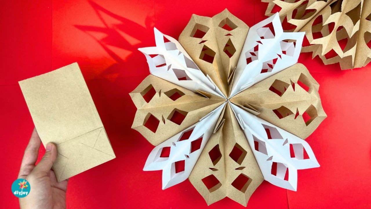 A hand holding a flat brown paper lunch bag beside a large fanned paper bag snowflake in brown and white