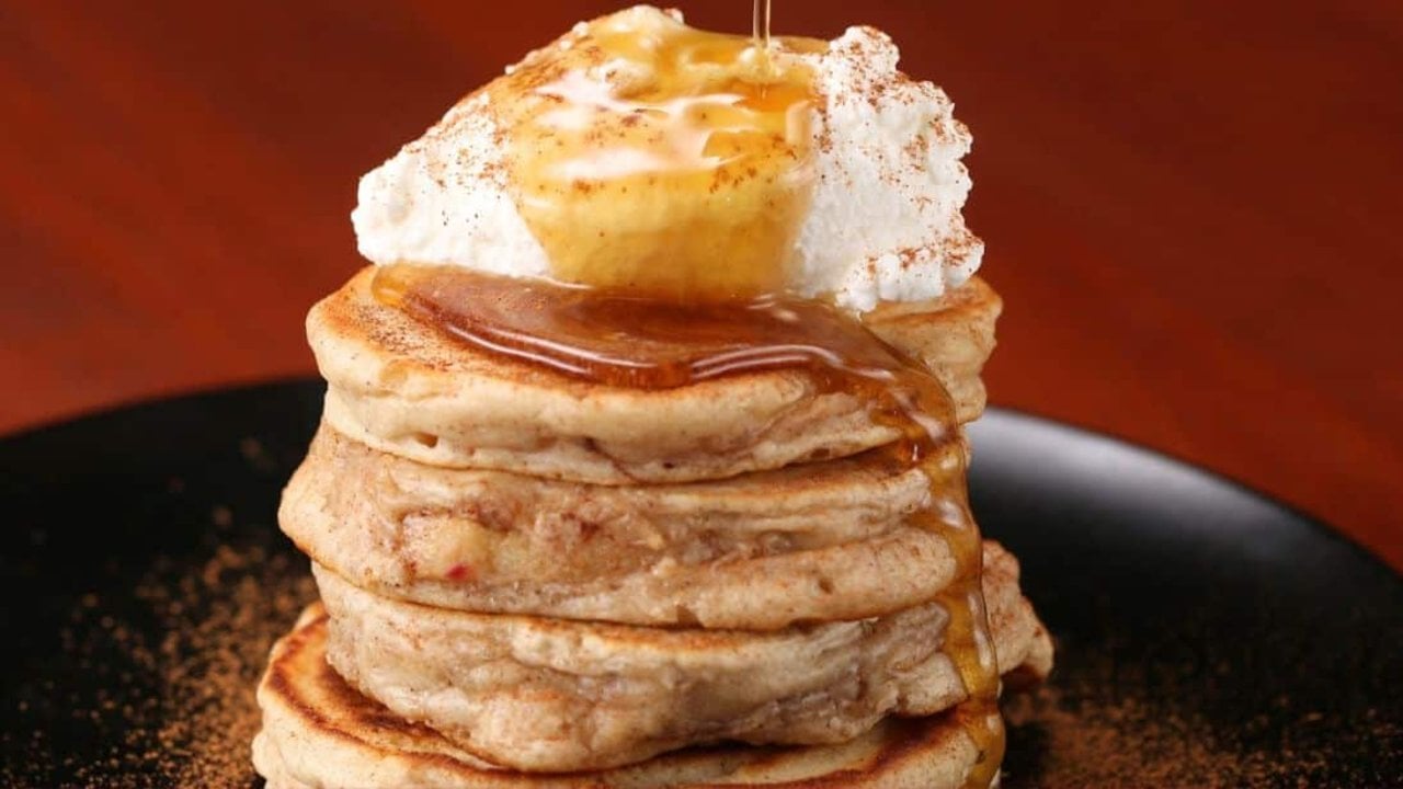 Apple ring pancakes stacked tall on a black plate topped with whipped cream and caramel syrup running down the side