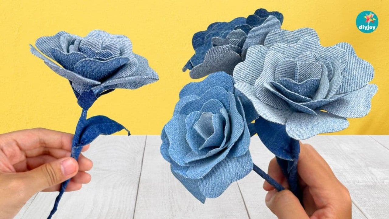 Denim roses in three shades of blue held in two hands against a bright yellow wall with more roses below