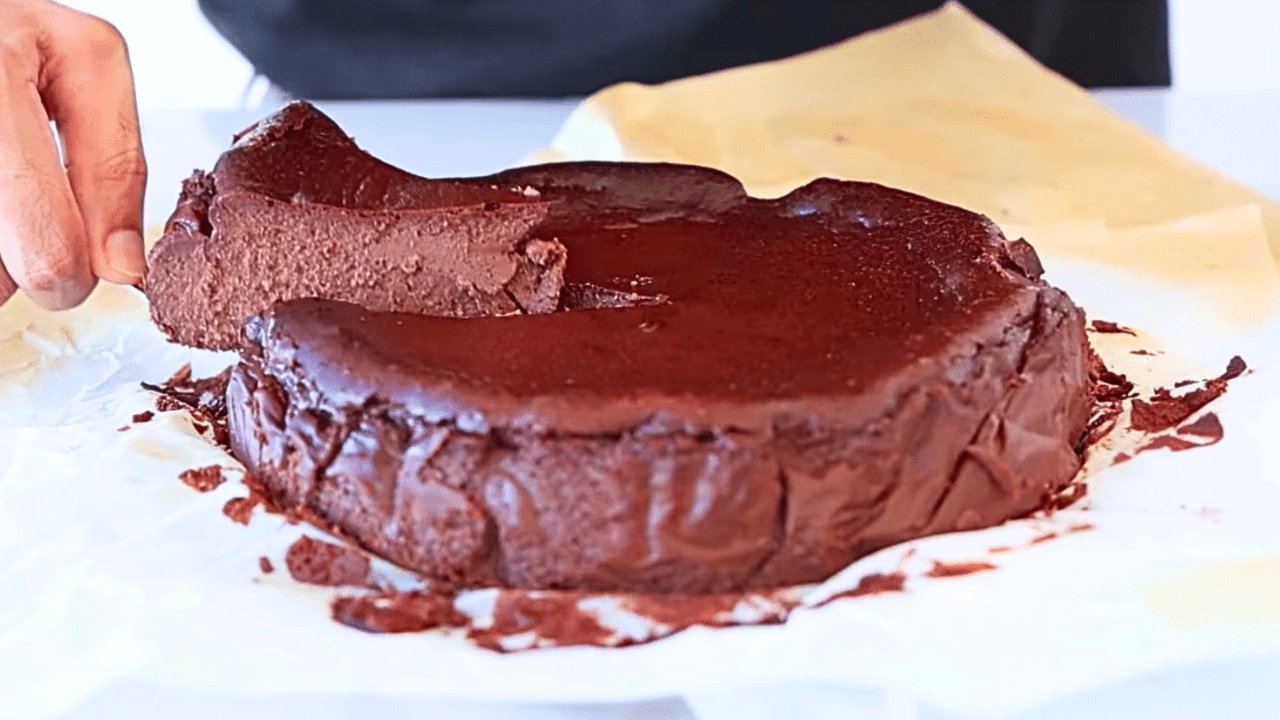 Whole chocolate basque cheesecake with a cracked deeply browned top on crumpled parchment, a hand steadying it