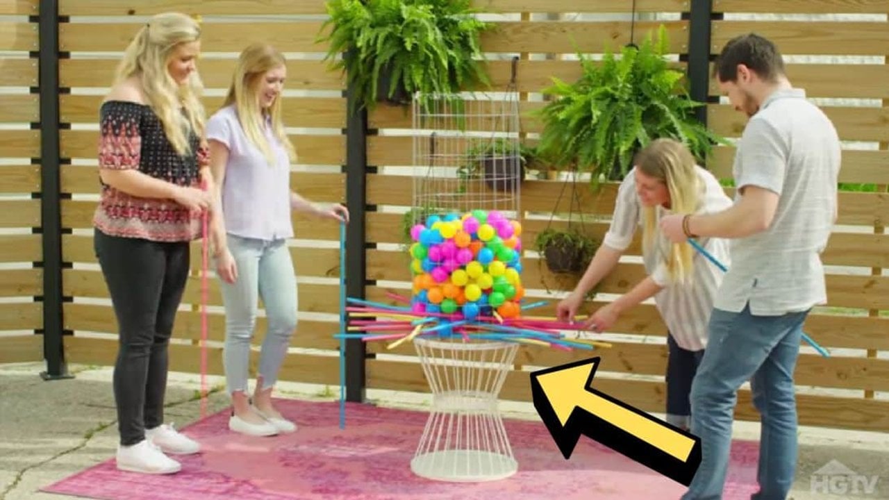 Four adults playing a giant DIY backyard kerplunk game, a wire tower of colorful balls held up on crossed dowels