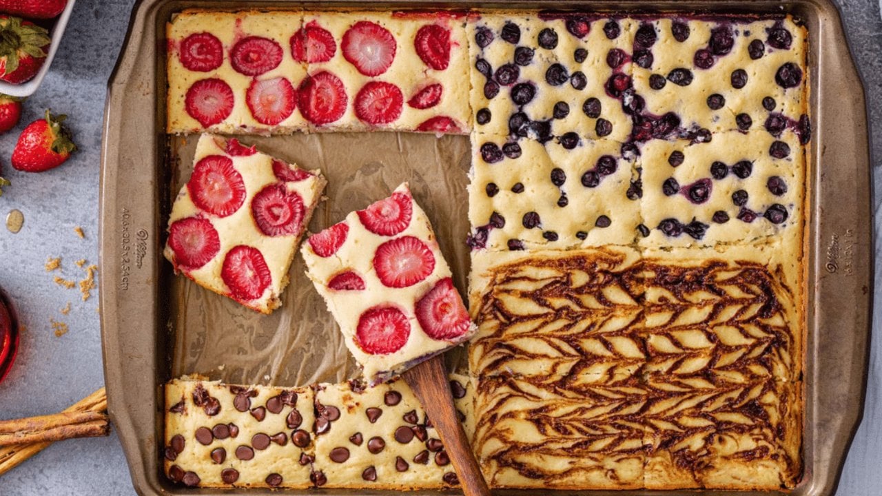 Sheet pan pancake cut into four quarters topped with strawberries, blueberries and chocolate, a spatula lifting a square