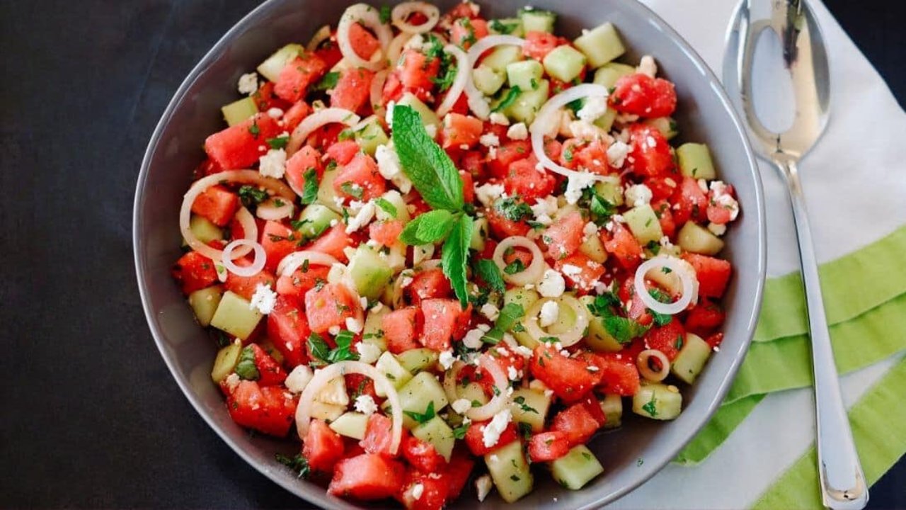 Watermelon salad with cubed melon, cucumber, thin sweet onion rings, mint and crumbled feta in a dark bowl