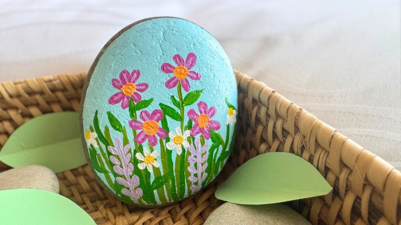 Smooth rock painted with pink and white flowers, green stems and purple lavender over a blue painted background