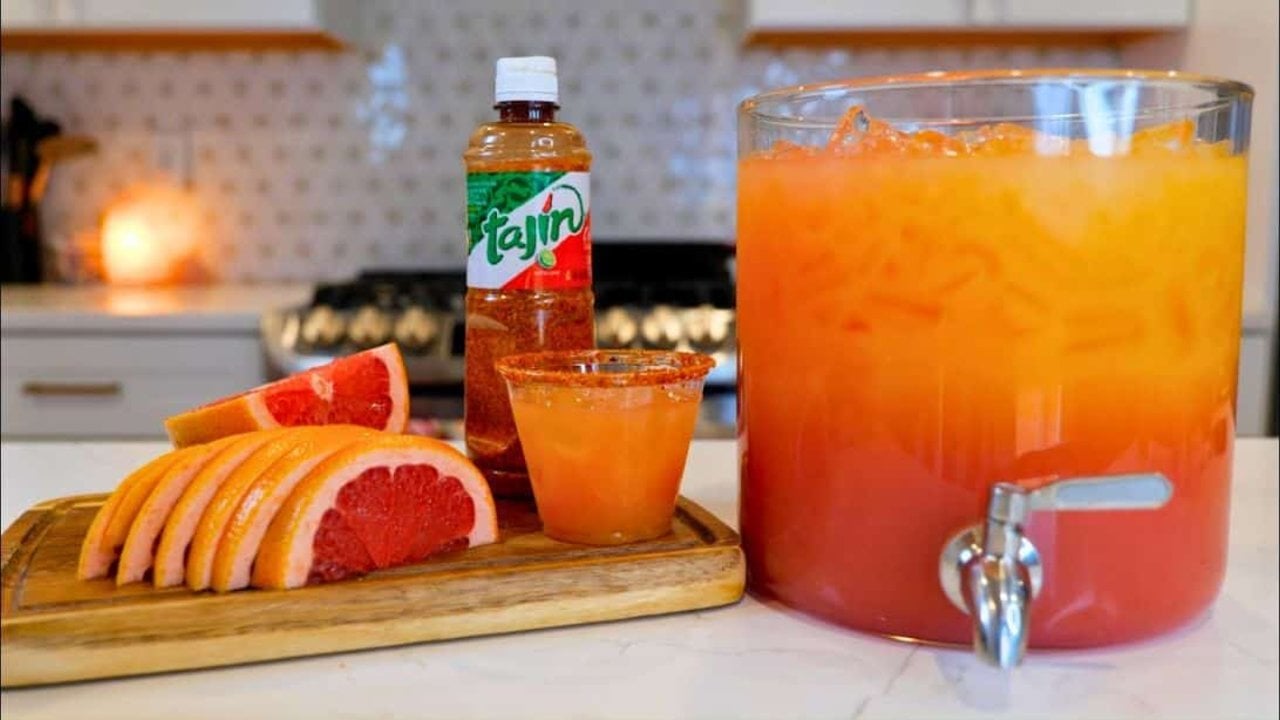 Grapefruit Cranberry Party Drink A glass beverage dispenser with a spigot beside one Tajin rimmed glass, a Tajin bottle and grapefruit slices