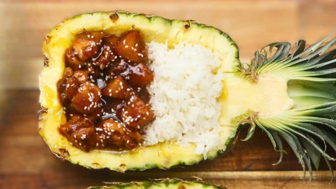 Sticky pineapple chicken served in a hollowed pineapple half with white rice and toasted sesame seeds