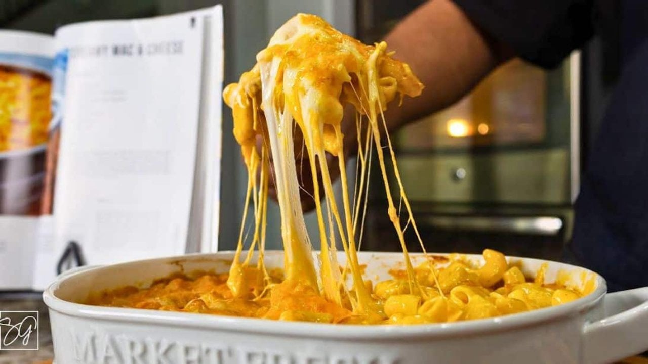 The Secret to Creamy Mac and Cheese A hand lifting a long cheese pull from a white oval dish of elbow macaroni, a recipe page propped behind