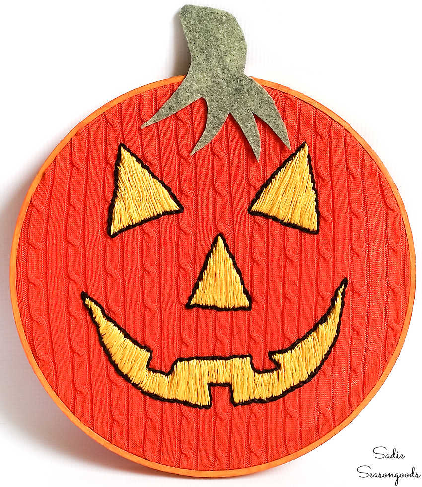Orange cable knit sweater in an embroidery hoop with a yellow jack-o-lantern face hand stitched onto it