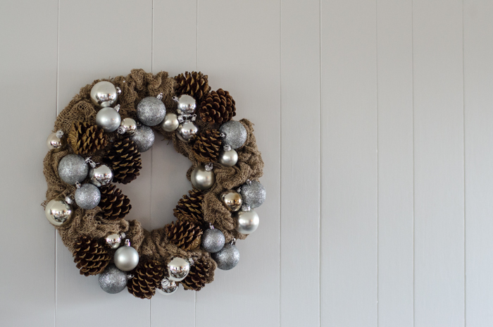 Burlap wreath studded with painted pinecones and silver baubles hanging on a shiplap wall for the holidays