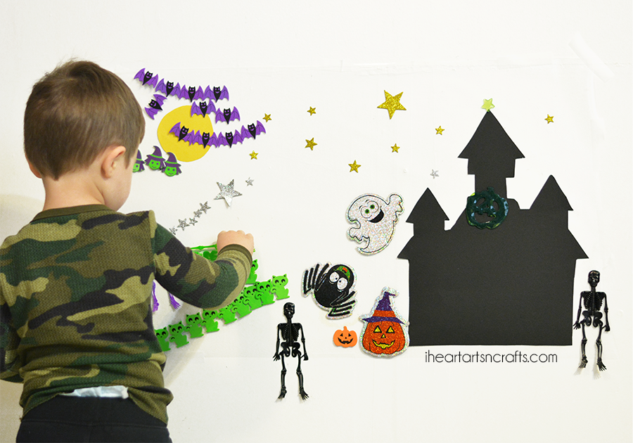 DIY Halloween sticky wall activity for kids, a boy sticking bat, ghost, haunted house and skeleton shapes onto contact paper taped to the wall
