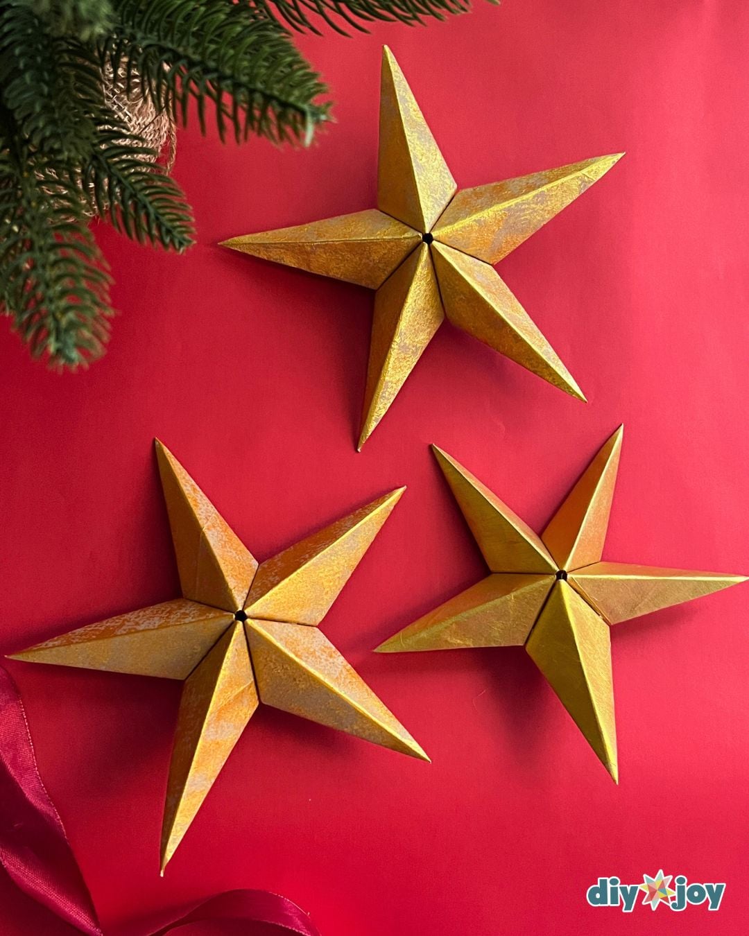 Paper bag star ornament - three finished 3D gold paper stars on a red background with a pine sprig, homemade decor
