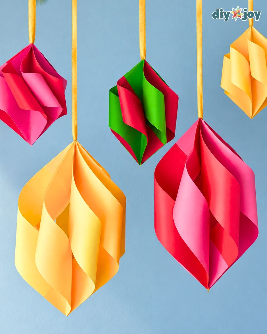 Paper Christmas ornaments in red, green, pink and yellow hang from gold ribbon, colorful DIY Christmas decor on a blue wall