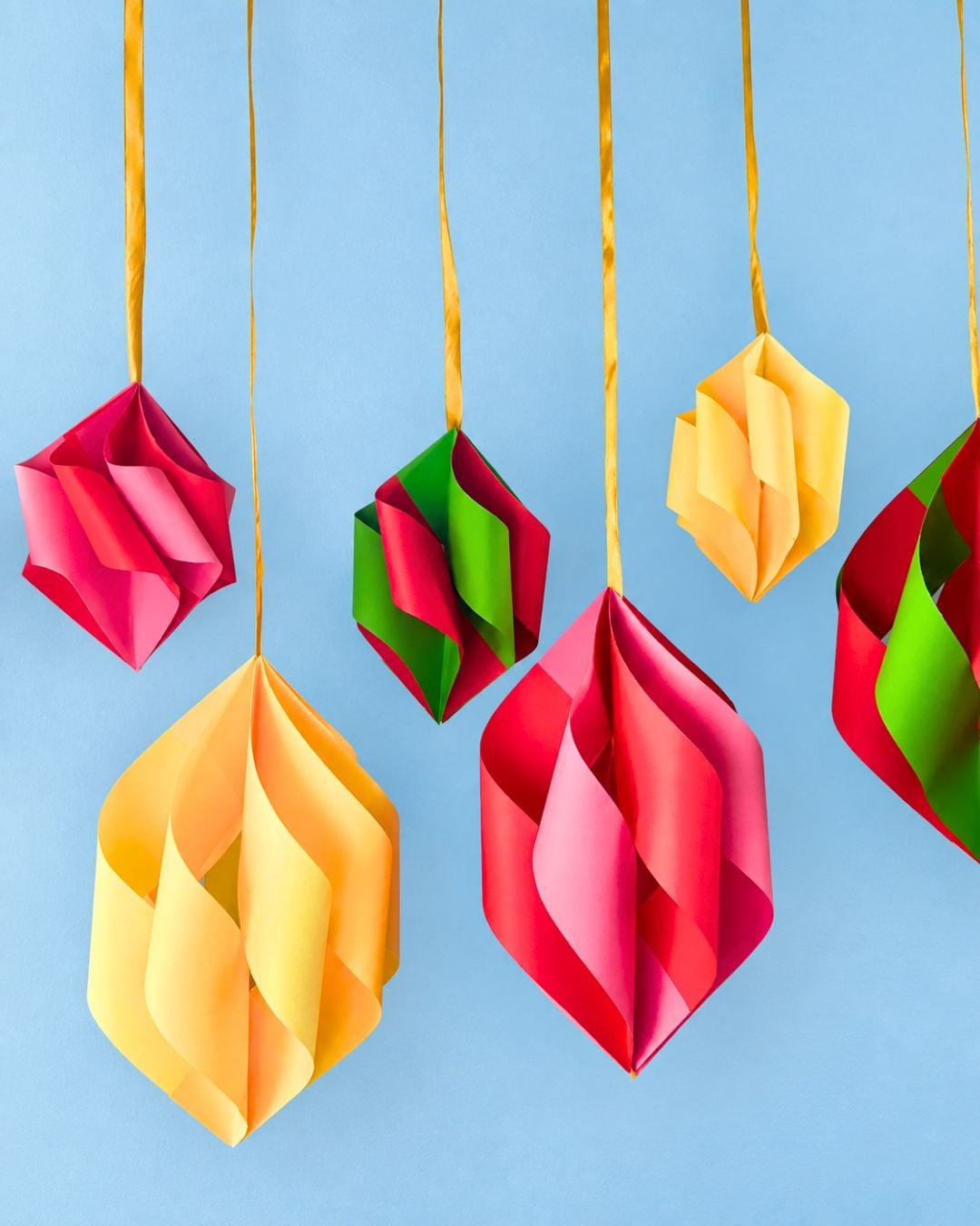 Pink, yellow, red and green paper ornaments hang on gold ribbons against a sky blue backdrop, fresh DIY Christmas decor