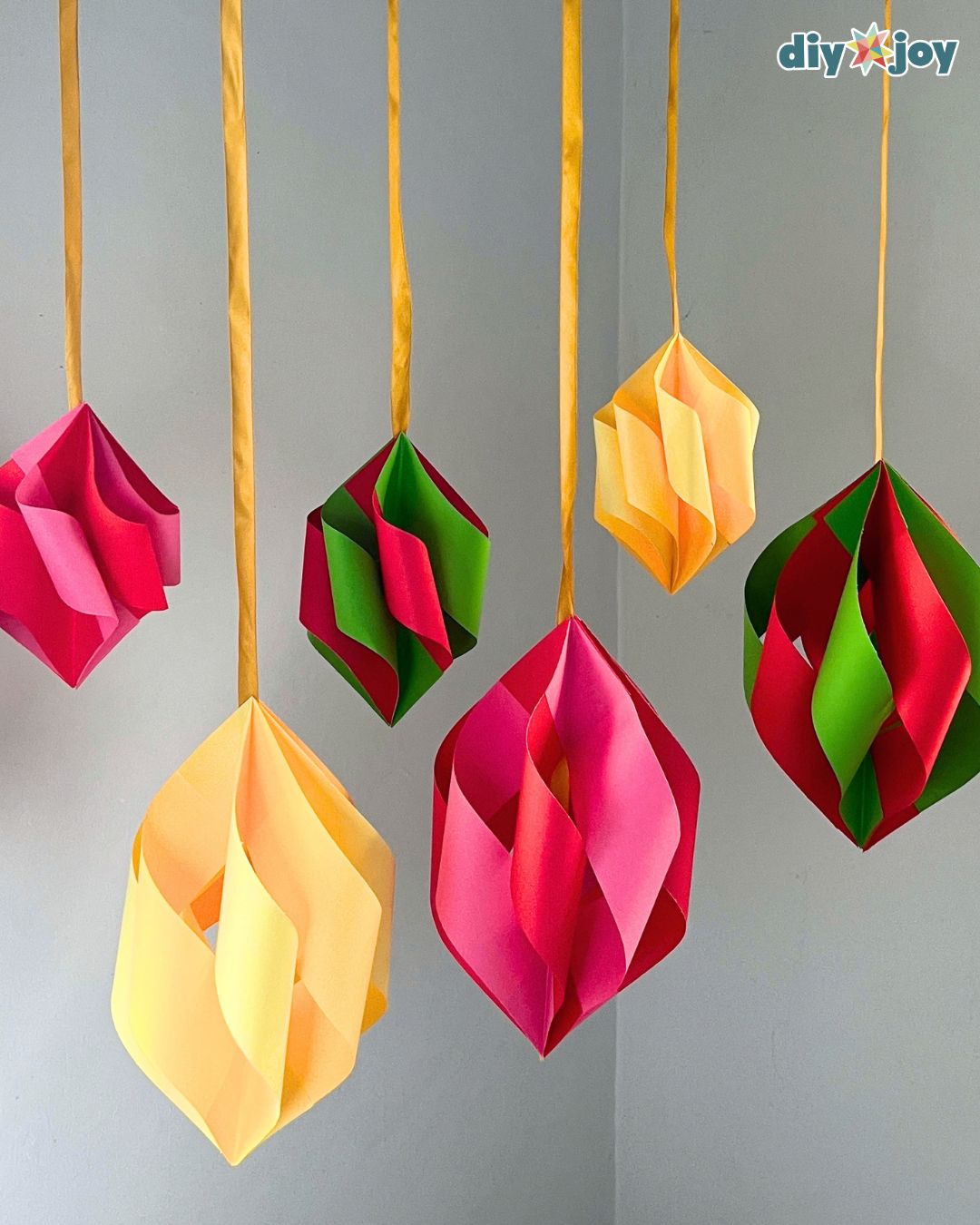 Seven folded paper ornaments hang at different heights in a white corner, easy DIY Christmas decor in festive colors