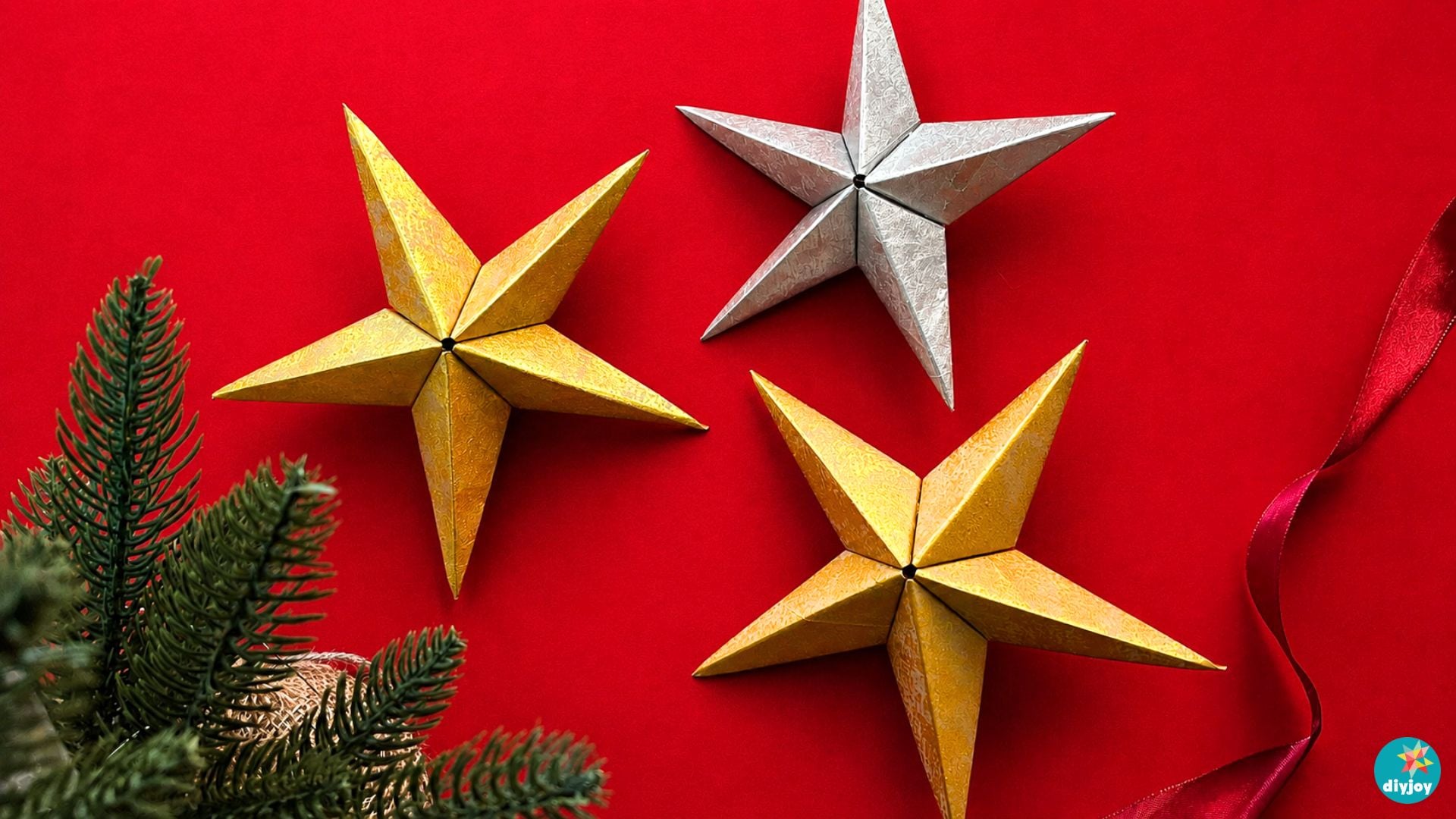 Paper Bag Star Ornament in Gold and Silver Three folded paper stars in gold and silver on a red background with pine sprigs and a length of satin ribbon