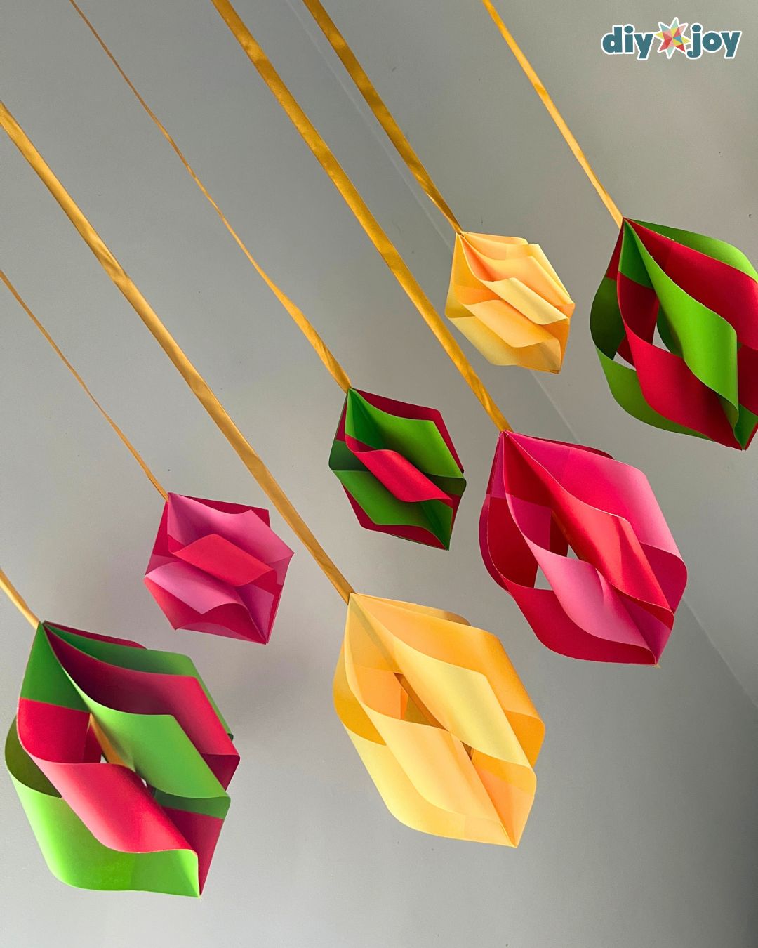 Angled view of colorful folded paper ornaments hanging on long gold ribbons, a five-minute DIY Christmas decor craft
