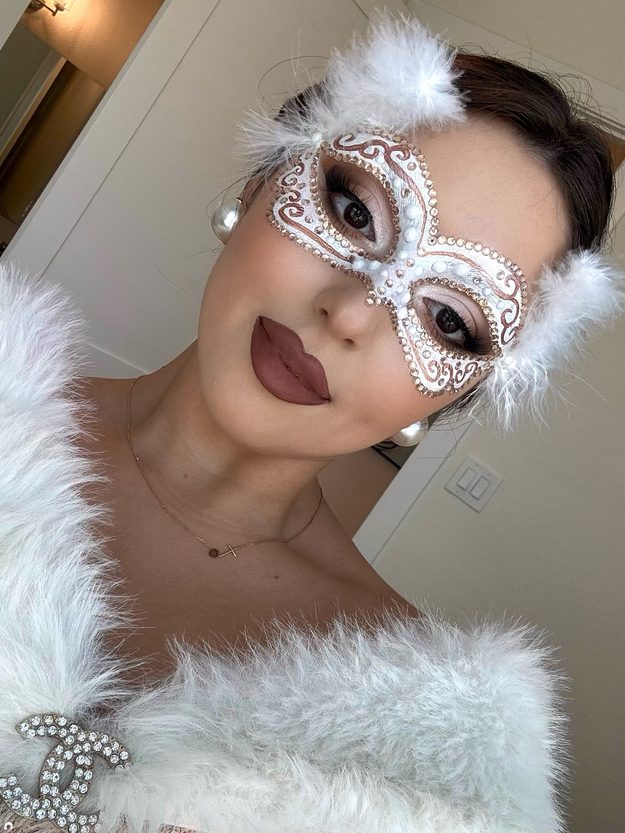 Masquerade Halloween makeup with a silver painted mask, rhinestone details, white feathers and dark lips
