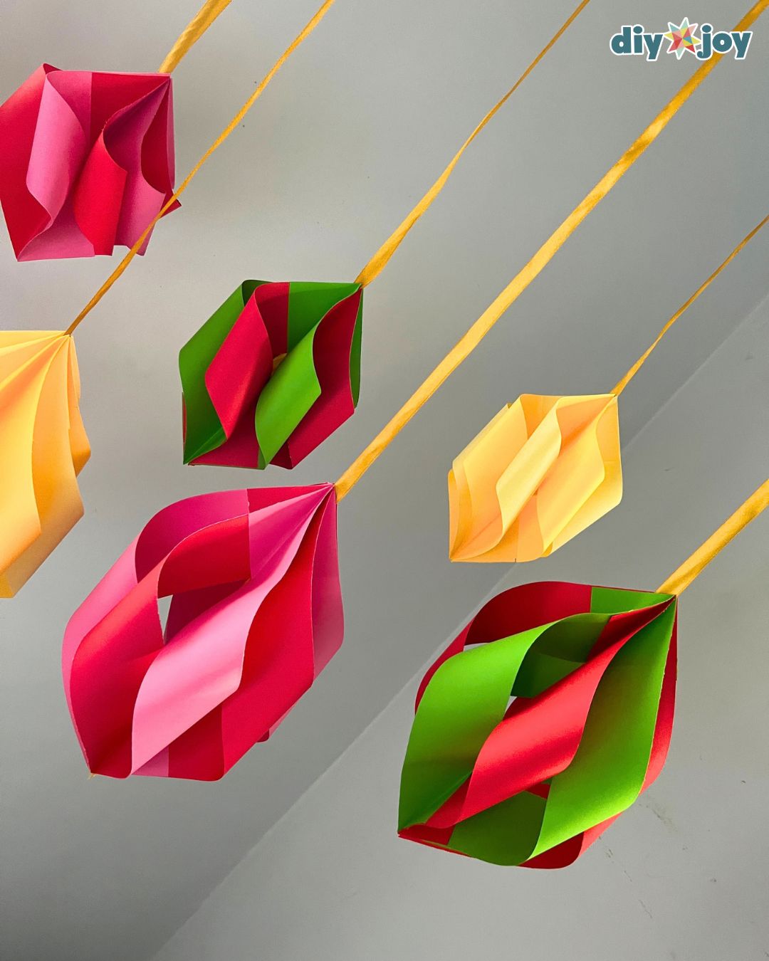 Red, pink, green and yellow paper ornaments float on gold ribbons against a gray wall, budget-friendly DIY Christmas decor