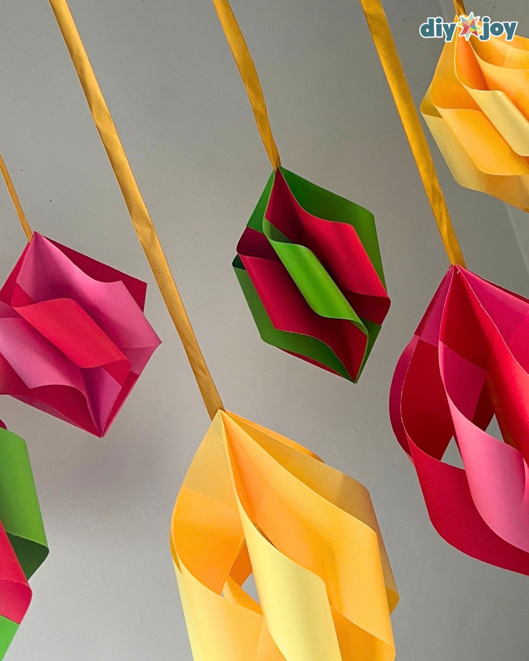 Close view of red, pink, green and yellow folded paper ornaments on gold ribbon, bright DIY Christmas decor for any room