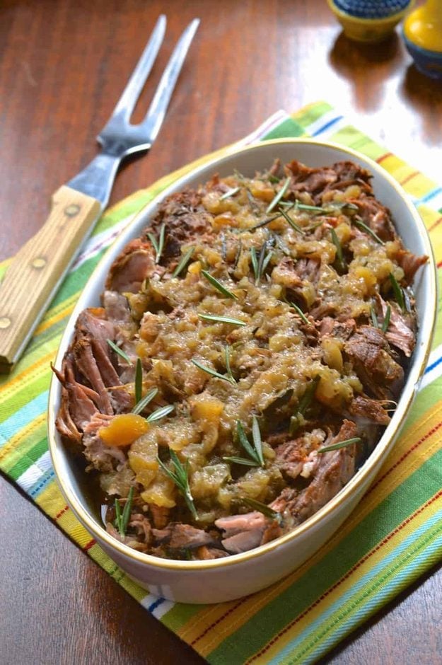 Apple Rosemary Slow Cooker Pork Roast Cooked pork roast under chunky apples and rosemary in a white oval dish beside a cloth and carving fork