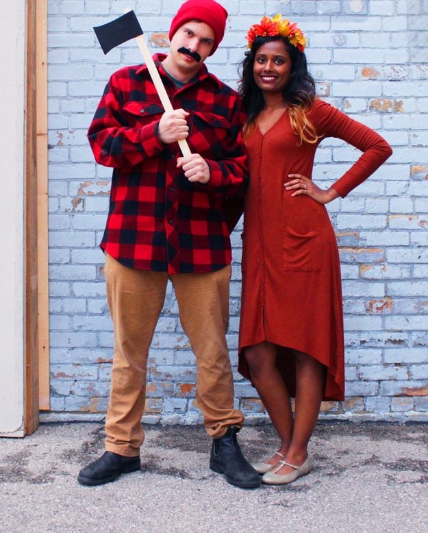 DIY Halloween couples costume, a lumberjack in red plaid holding a foam axe beside a tree in a rust dress and leaf crown