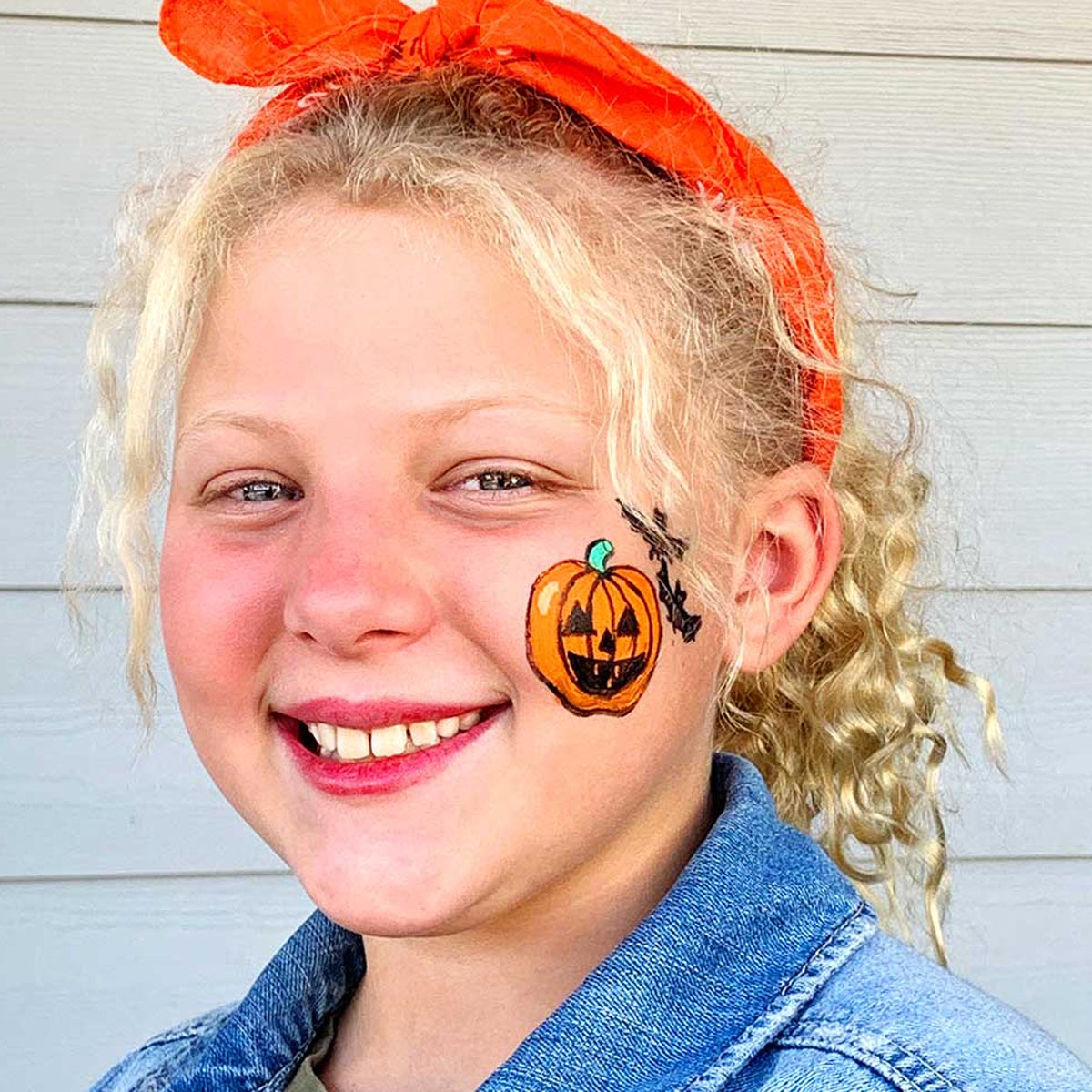 Easy Halloween face paint with a small orange pumpkin and black bat design painted on a smiling girl's cheek