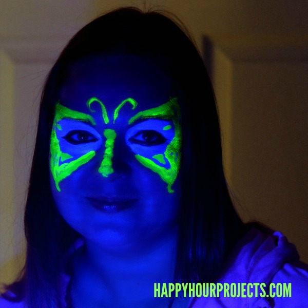 Blacklight butterfly face paint glowing neon green and blue around the eyes against bright blue skin