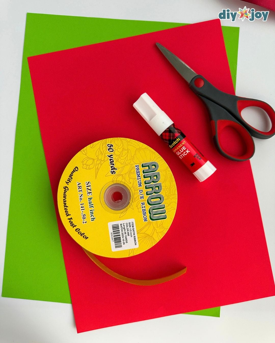Supplies for DIY Christmas decor laid out: red and green paper, scissors, a glue stick and a spool of gold ribbon