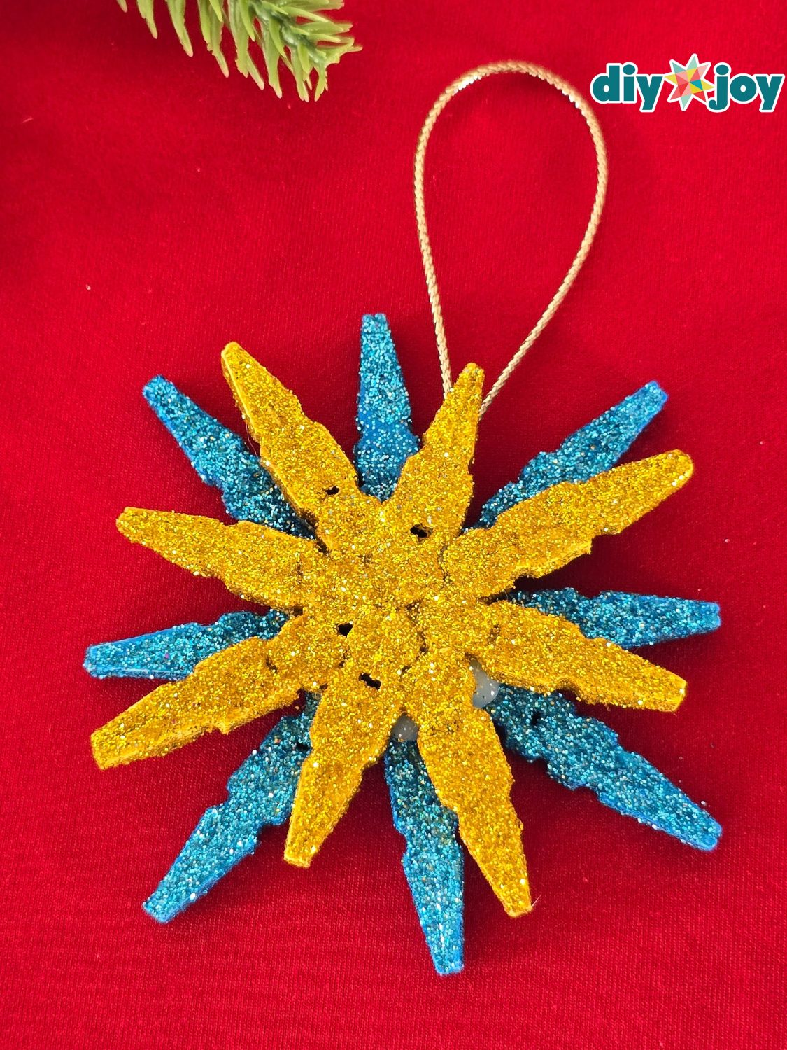 Glitter Clothespin Snowflake Ornament Snowflake ornament made from glittered clothespin halves in gold layered over blue, with a gold cord loop on red fabric