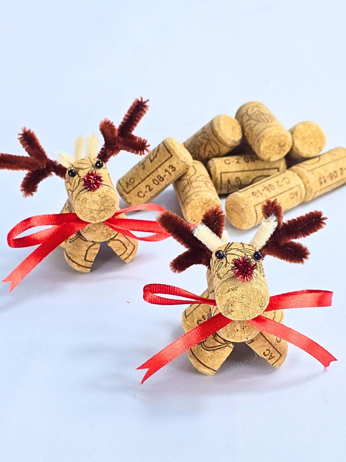 Wine Cork Reindeer Christmas Craft Two wine cork reindeer with brown pipe cleaner antlers, red pom pom noses and red ribbon scarves beside a pile of corks