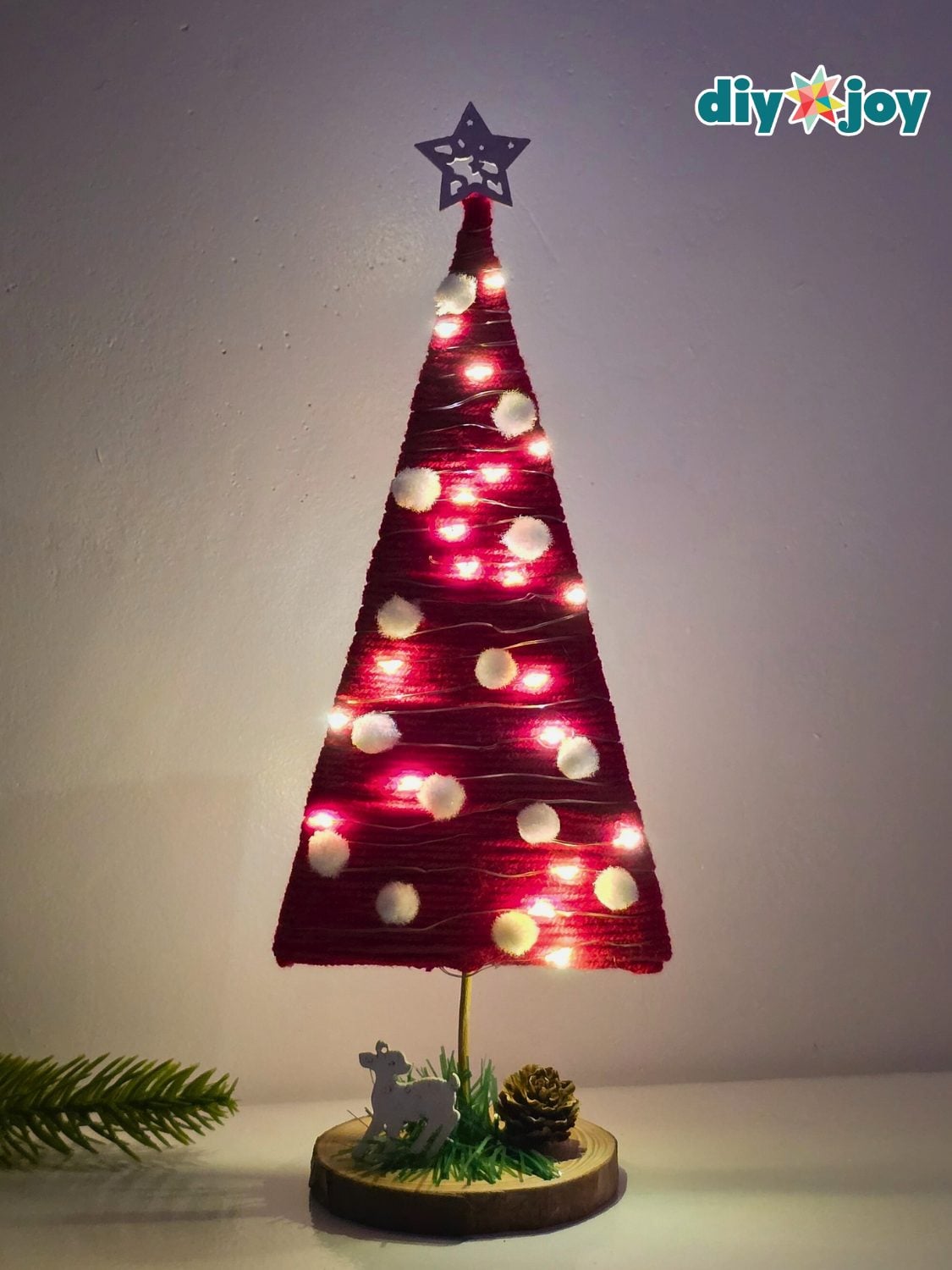 Yarn Christmas Tree With Fairy Lights Red chunky yarn Christmas tree wrapped on a cardboard cone, lit with fairy lights and dotted with white pom poms
