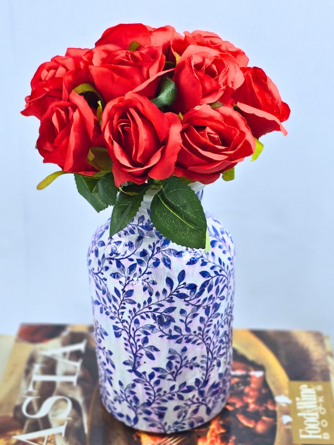 Glass jar decoupaged in a blue and white floral napkin pattern holding a bunch of red roses beside cookbooks