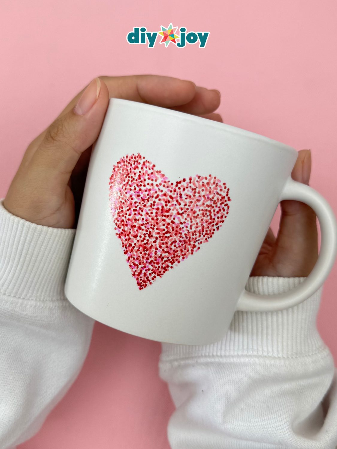White mug with a heart outlined in red and pink marker dots held in two hands against a pink background