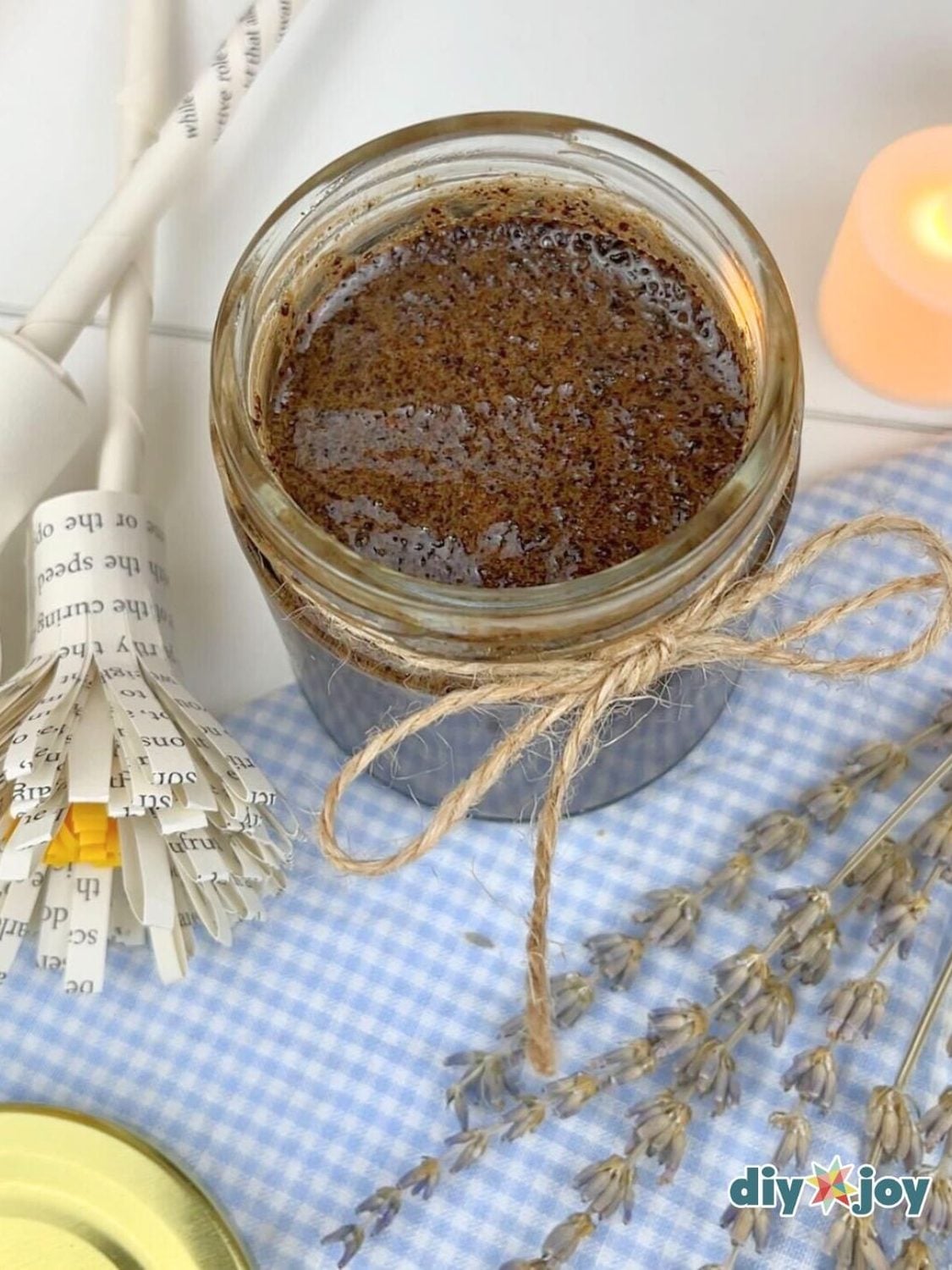 Glass jar of dark brown coffee body scrub with a wooden spoon resting in it on a pale marble counter